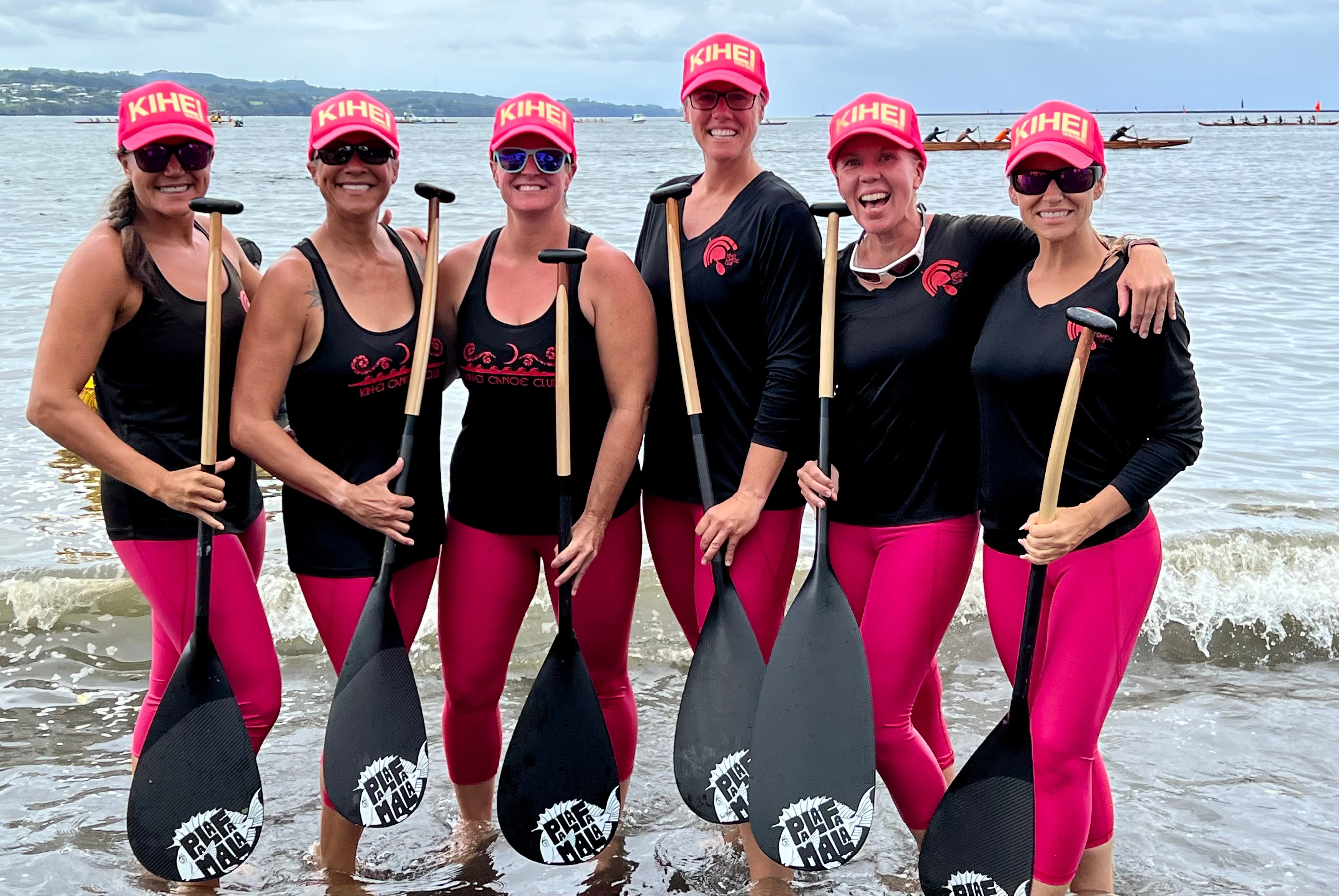 Group of women in black shirts and pink pants with paddle sports equipment on a beach.